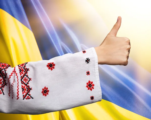 Woman in Ukrainian clothes shows symbol ok against Ukrainian fla — Stock fotografie
