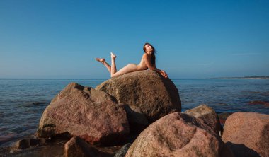 Genç bir kadın açık mavi gökyüzünün altında deniz kenarındaki büyük bir kayanın üzerinde çıplak poz veriyor, güneşin ve okyanus esintisinin tadını çıkarıyor.