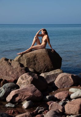 Geniş bir kayanın üzerinde çıplak poz veren genç bir kadın, arka planda deniz ve gökyüzü, sakin ve doğal bir sahne yaratıyor.