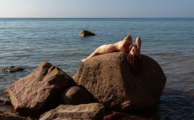 Geniş bir kayanın üzerinde çıplak poz veren genç bir kadın. Arka planda okyanus ve gökyüzü. Huzurlu ve doğal bir manzara yaratıyor.