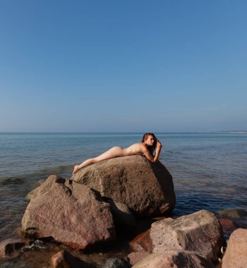 Genç bir kadın güneşli bir günde deniz kenarındaki büyük bir kayanın üzerinde çıplak poz veriyor.
