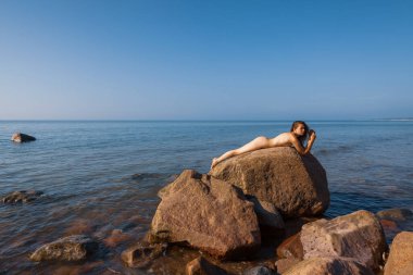 Genç bir kadın deniz kenarındaki büyük bir kayanın üzerinde çıplak poz veriyor. Güneşin ve manzaranın tadını çıkarıyor.