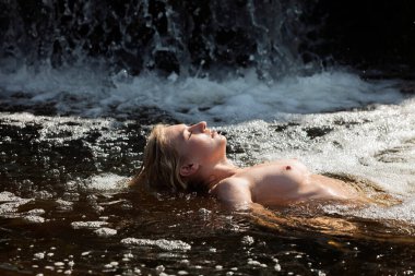 Nehirde çıplak poz veren genç bir kadın, serin suyun ve sıcak güneşin tadını çıkarıyor.