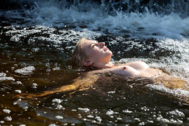 Çıplak poz veren genç bir kadın, serin suyun ve güneş ışığının tadını çıkarıyor.