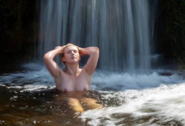 Huzurlu bir şelalede çıplak poz veren genç bir kadın doğanın güzelliğini ve o anın sükunetini kucaklıyor.