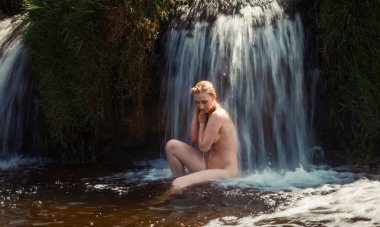 Çıplak poz veren genç bir kadın şelalenin altında oturmuş, serin suyun ve doğanın güzelliğinin tadını çıkarıyor.