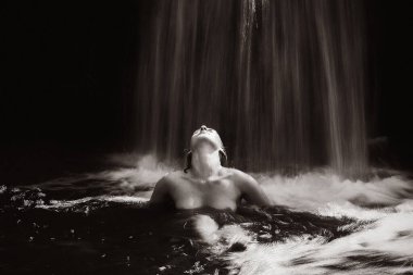 Çıplak poz veren genç bir kadın sanatsal monokrom çekiminde serinletici suyun üzerine düşmesinden hoşlanıyor.
