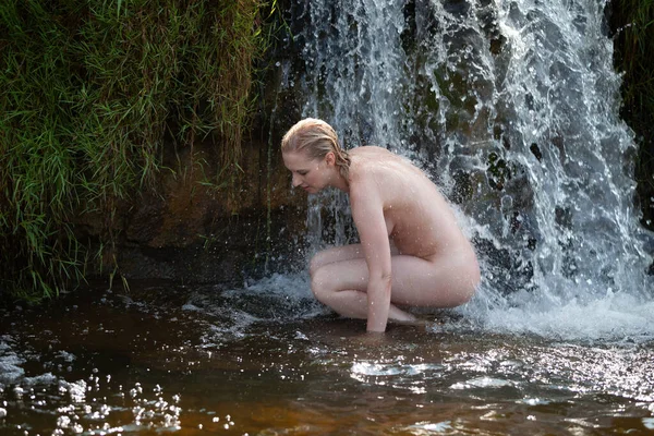 Doğal ortamında çıplak poz veren genç bir kadın, şelalenin ferahlatıcı suyunun tadını çıkarıyor.