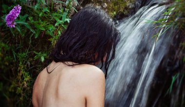 Genç kadın çıplak poz veriyor, arkası ıslak koyu saçlı, yemyeşil yaprakların ve canlı mor bir çiçeğin ortasında akan bir şelalenin yanında duruyor. Cildinde su damlacıkları görünüyor..