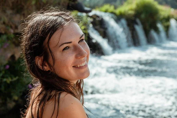 Genç kadın çıplak poz veriyor, ıslak saçları yüzünü çerçeveliyor ve gülümsüyor ve kameradan başka tarafa bakıyor. Arka planda bulanık doğal bir şelale ve akan bir su var..