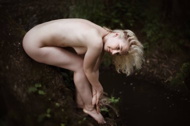 Kısa kıvırcık saçlı genç bir kadın karanlık bir yosunlu ağaç gövdesinde çıplak halde ayak bileklerini tutarken ve loş ormanda yavaşça kameraya bakarken..