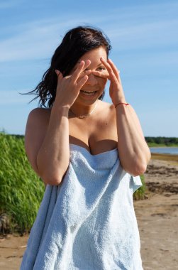 Koyu saçlı bir kadın, açık mavi bir havluya sarılmış göğsünü örtüyor, açık havada uzun yeşil kamışların yanında duruyor ve açık mavi gökyüzünün altında kumlu bir sahil..