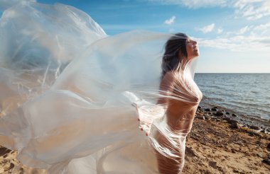 Rüzgarlı deniz sahilinde, plastik ambalajın arkasında güzel, çıplak bir kadın. Polietilen film altında sanatsal vücut portresi, doğa arka planı. Kavramsal açık hava fotoğrafı.