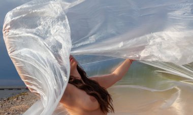 Fine art nude woman behind plastic wrap on windy sea beach. Artistic body portrait under polyethylene film, nature background. Conceptual outdoor photo.
