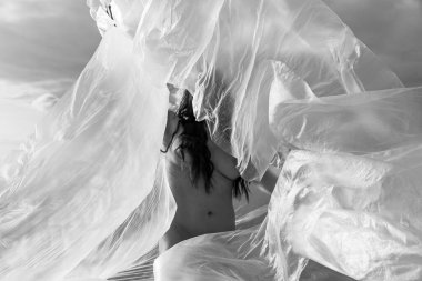 Rüzgarlı deniz sahilinde, plastik ambalajın arkasında güzel, çıplak bir kadın. Polietilen film altında sanatsal vücut portresi, doğa arka planı. Kavramsal açık hava fotoğrafı.