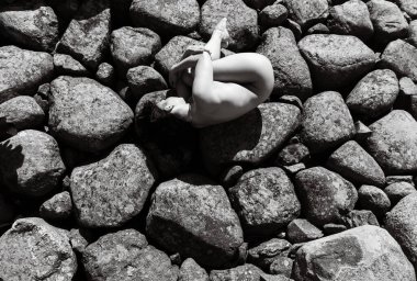 Siyah-beyaz fotoğrafçılık, çıplak genç bir kadının cenin pozisyonunda kıvrıldığını gösteriyor. Büyük, pürüzlü, dokulu kayaların üzerinde..