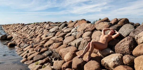 Çıplak genç bir kadın, parlak mavi bir gökyüzünün altında, ince bulutlu, büyük, dalgalı bir dalgakıranın üzerinde uzanır..