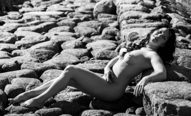Çıplak bir kadın dalgakıranın büyük, pürüzlü taşlarına yaslanır. Güneş ışığı, vücudun kıvrımlarını dokulu kaya yüzeyine vurgular. Siyah-beyaz fotoğrafçılık.