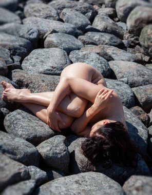 Çıplak, yetişkin bir dişi parlak güneş ışığı altında büyük gri taşların üzerinde yatıyor. Koyu renk saçlar kafanın altındaki kayaların üzerinde duruyor. Deri pürüzsüz ve bronzlaşmış görünüyor.