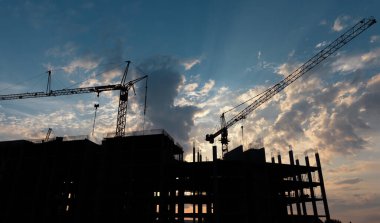 İnşaat halindeki büyük bir binanın siluetleri ve dramatik bir gökyüzüne karşı ön planda birden fazla vinç var..