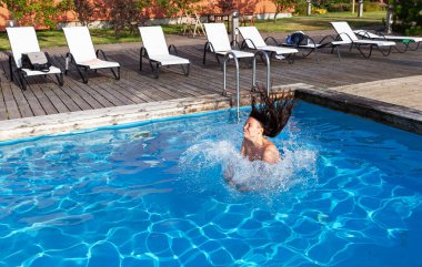 A woman emerges from bright blue water creating a large splash. White lounge chairs sit on a wooden deck beside the pool. Green foliage is visible in the background.
