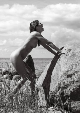 Beyaz çıplak bir kadın çimenli bir sahilde büyük bir kayaya yaslanıyor. Kadın bulutlu bir gökyüzünün altında açık bir ağızla başını arkaya eğiyor. Siyah-beyaz fotoğrafçılık.