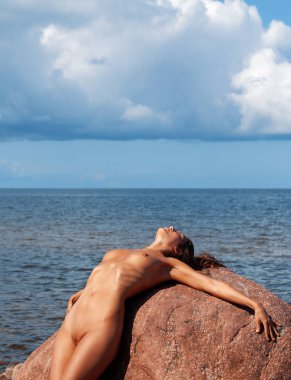 Çıplak bir kadın deniz kenarındaki büyük kırmızı bir kayaya yaslanıyor. Güneş ışığı gövdeyi ve yüzü aydınlatırken kümülüs bulutları ufuktaki mavi gökyüzünü doldurur..