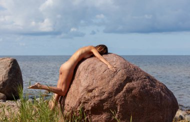 Genç çıplak bir kadın çimenli bir kıyı şeridindeki büyük kırmızı bir kayanın üzerine eğiliyor. Deniz bulutlu mavi bir gökyüzünün altında ufka kadar uzanıyor. Güneş ışığı kadının sırtını aydınlatır..