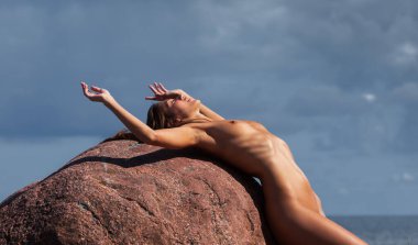 Genç, narin bir kadın, bulutlu mavi bir gökyüzünün altında denizin yakınındaki büyük, kırmızımsı bir taşın üzerinde çıplak yatıyor. Kadın geriye doğru eğiliyor, kollarını havaya kaldırarak..