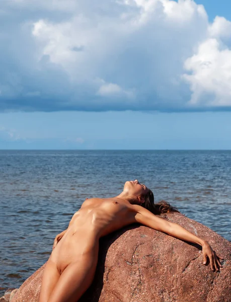 Çıplak bir kadın deniz kenarındaki büyük kırmızı bir kayaya yaslanıyor. Güneş ışığı gövdeyi ve yüzü aydınlatırken kümülüs bulutları ufuktaki mavi gökyüzünü doldurur..