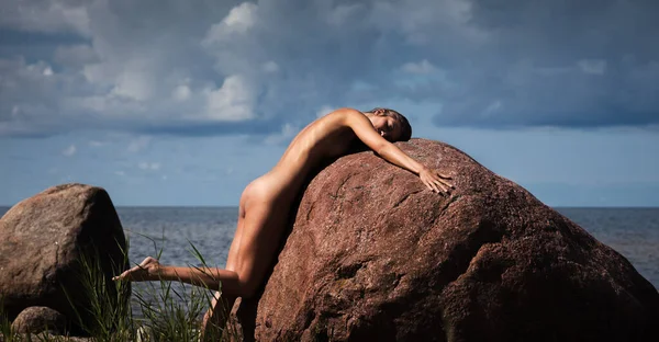 Genç, narin bir kadın, bulutlu bir gökyüzünün altında, deniz kenarındaki büyük, kırmızımsı kahverengi bir kayanın üzerinde yatıyor. Vücut, karanlık suya ve dramatik bulutlara karşı güneş ışığıyla aydınlanır..