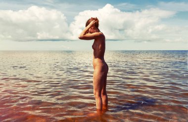 Çıplak bir kadın bulutlu bir gökyüzünün altında denizin kenarında sığ sularda duruyor. Kadının vücudunu kumla kaplamış ve iki elini de saçına kaldırmış..