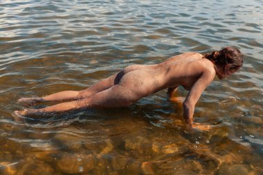 Çıplak bir kadın sığ sularda, arkası ve bacakları kum kaplı şınav çeker. Kadın figürü, el ve ayak parmakları üzerinde dengede durur..