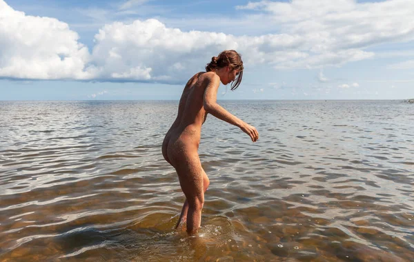 Genç çıplak bir dişi bulutlu mavi bir gökyüzünün altında, kıyıya yakın sığ ve bulanık bir suda duruyor. Kum kadın vücudunu kaplar..