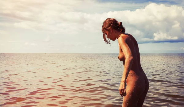 Genç çıplak bir kadın bulutlu bir gökyüzünün altında kıyı şeridine yakın bir yerde kısmen suyun altında duruyor. Kadının vücudu kumla kaplıdır ve kadın aşağıya, suya bakar..