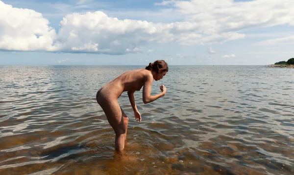 Genç çıplak bir kadın kıyı şeridine yakın sığ ve dalgalı bir suda eğilmiş duruyor. Kum kadının sırtını ve bacaklarını kaplıyor. Ufuk suyun gökyüzüne vuruşunu gösteriyor. Başında büyük beyaz bulutlar..