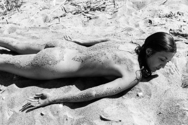 Çıplak bir kadın kumlu bir plajda uzanmış sırtı, kalçaları ve kolları tahıllarla kaplı. Sert güneş ışığı figürün etrafında derin gölgeler oluşturur. Siyah-beyaz fotoğrafçılık.