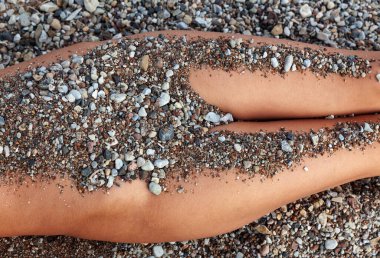 Bronzlaşmış bacaklar arka planda karışık çakıl taşlarına ve kaba kuma karşı yatay olarak dinleniyor. Küçük, çok tonlu taşlar deriye iki farklı yatay bantta tutunurlar..