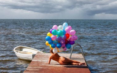 Çıplak bir figür, ağır bulutlu bir gökyüzünün altındaki karanlık ve dalgalı suya uzanan ahşap bir rıhtımda yatar. Büyük bir demet parlak renkli balon figürün sırtında tutulur..