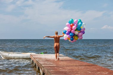 Kısa sarı saçlı çıplak bir kadın, koyu suya uzanan ahşap bir iskelede duruyor, elinde bir sürü renkli balon tutuyor. Gökyüzü açık mavi ve dağınık beyaz bulutlar.