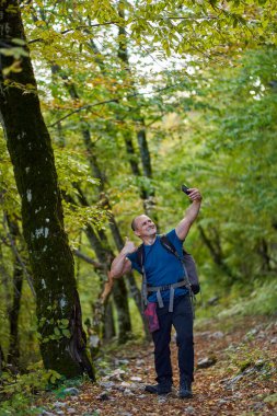 Sırt çantalı bir adam ormanda yürürken selfie çekiyor.
