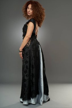 50 yaşında güzellik. Uzun elbiseli, kıvırcık saçlı olgun bir kadının stüdyo portresi.