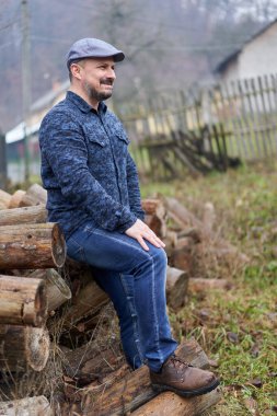Kot pantolonlu, gömlekli ve şapkalı çiftçi kış için kesilecek kütüklerin üzerinde oturuyor.
