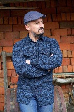 Mavi gömlekli ve kepli sakallı adam bir yığın tuğlanın yanında.