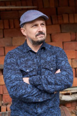 Mavi gömlekli ve kepli sakallı adam bir yığın tuğlanın yanında.