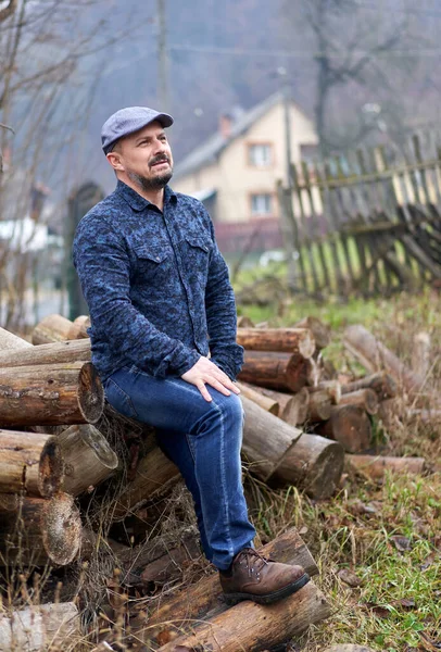 冬のために切り刻まれる丸太の山に座っている青いジーンズ シャツ 帽子の農家 — ストック写真