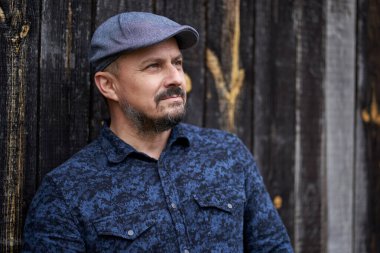 Mavi gömlekli ve kepli orta yaşlı bir adamın ahır ahşap arka planına yaklaşması.