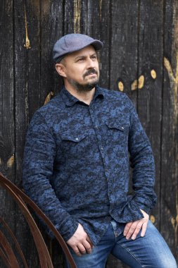 Mavi gömlekli ve kepli orta yaşlı bir adamın ahır ahşap arka planına yaklaşması.