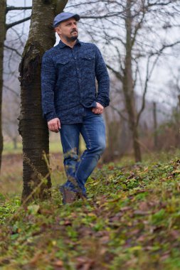 Bahçedeki bir ağaca yaslanmış kot pantolonlu ve gömlekli çiftçi.