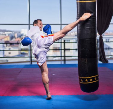 Kickboks dövüşçüsü spor salonunda kum torbasına vuruyor, yarışma için antrenman yapıyor.